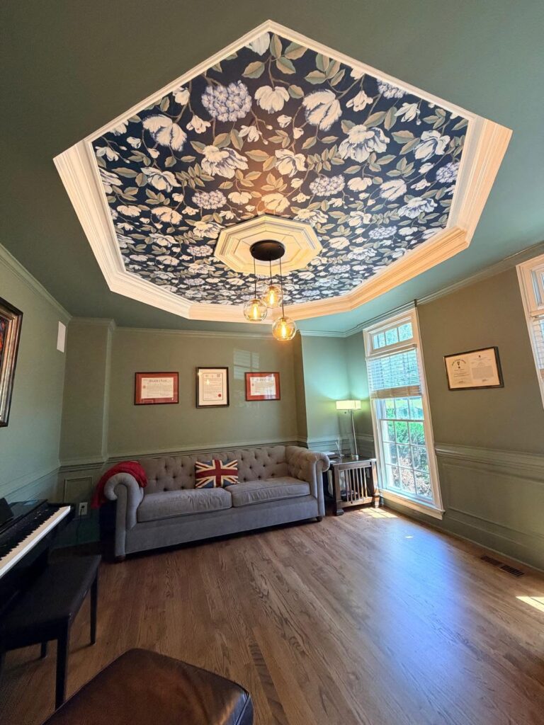 Intentional home design is featured in this music room with a piano, couch, and windows bringing light into a green room with floral wallpaper on the ceiling.