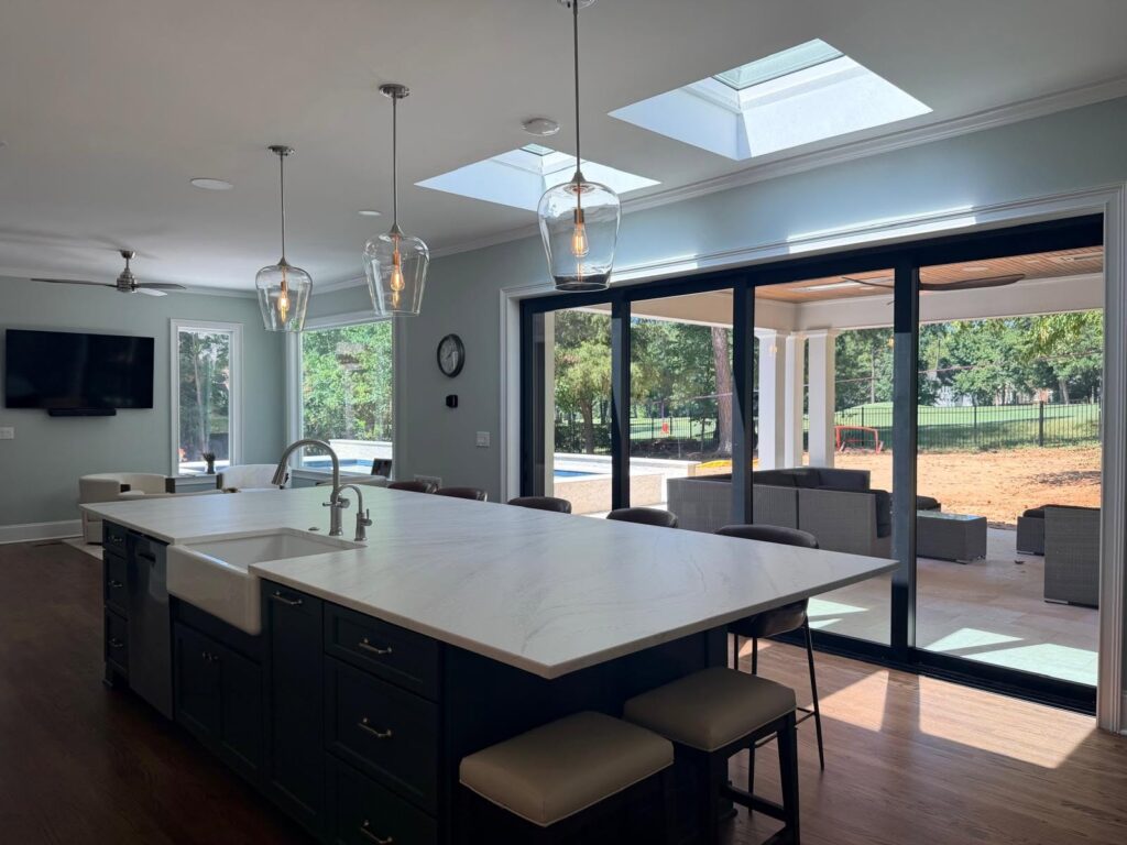 view from inside the enclosed living area looking on the modern pool. A large island is central with pendant lights overhead, bar stools behind and large windows with view to the backyard.