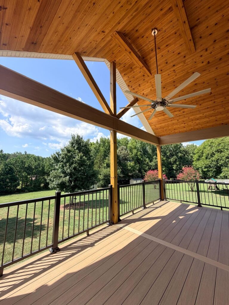 Timeless home design seen in this large porch addition with wood ceiling, huge ceiling fan and open railings looking out over a green backyard.